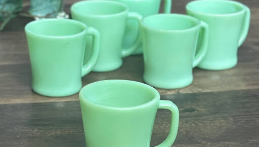 A group of six vintage Fire-King Jadeite D-handle mugs arranged on a wooden surface. Though they appear identical, slight differences in base design can affect their value.