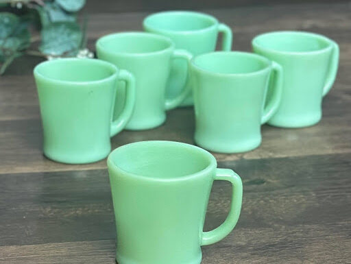 A group of six vintage Fire-King Jadeite D-handle mugs arranged on a wooden surface. Though they appear identical, slight differences in base design can affect their value.